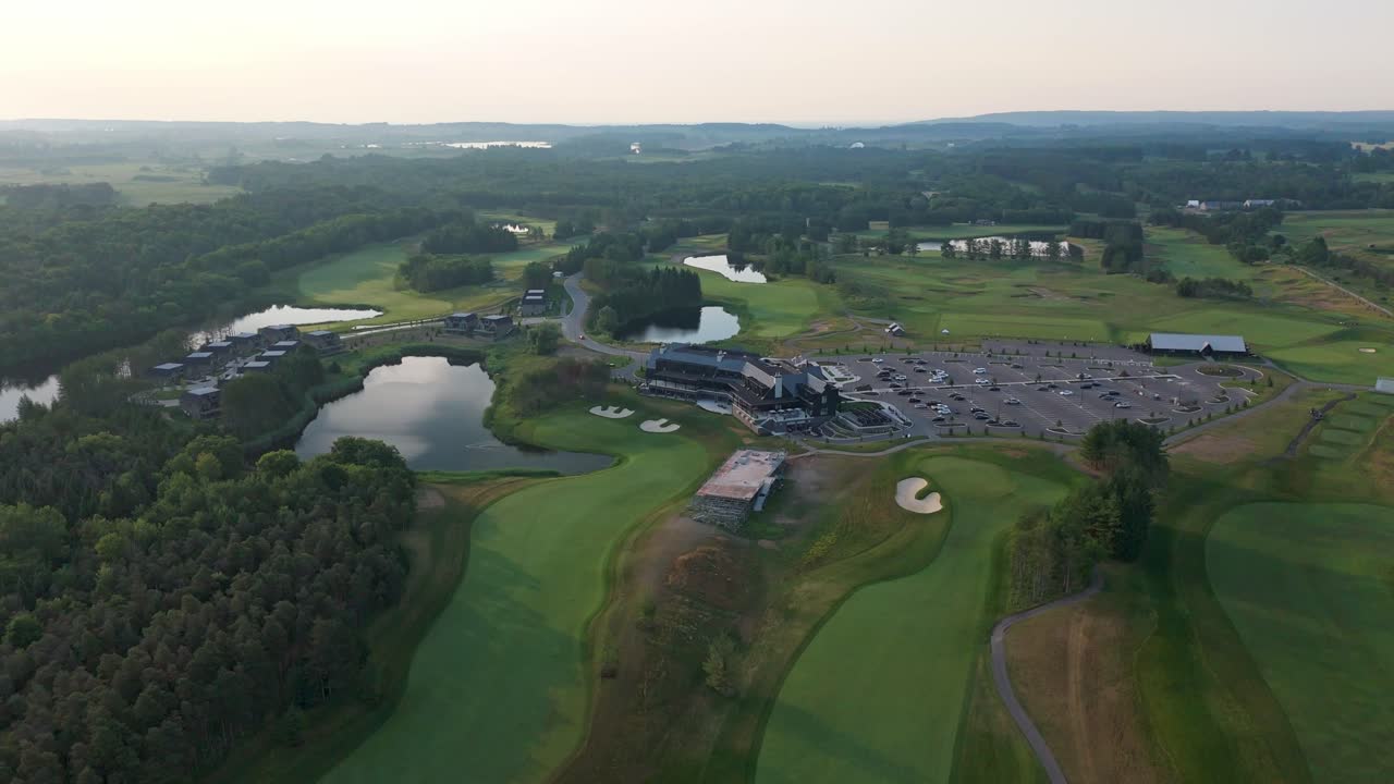 Descending Aerial Shot Of Lush Fairways And Clubhouse During Sunrise At A Golf Course