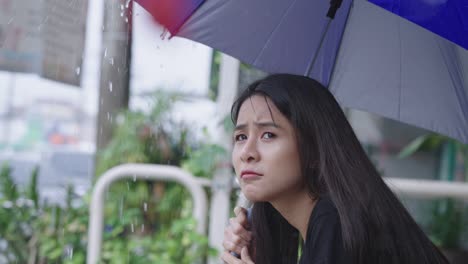 young adult asian female holding umbrella sit down on the street side walk on raining day, rainy season, feeling disappointed stuck in the rain with frustration anxiety, waiting for the rain to stop