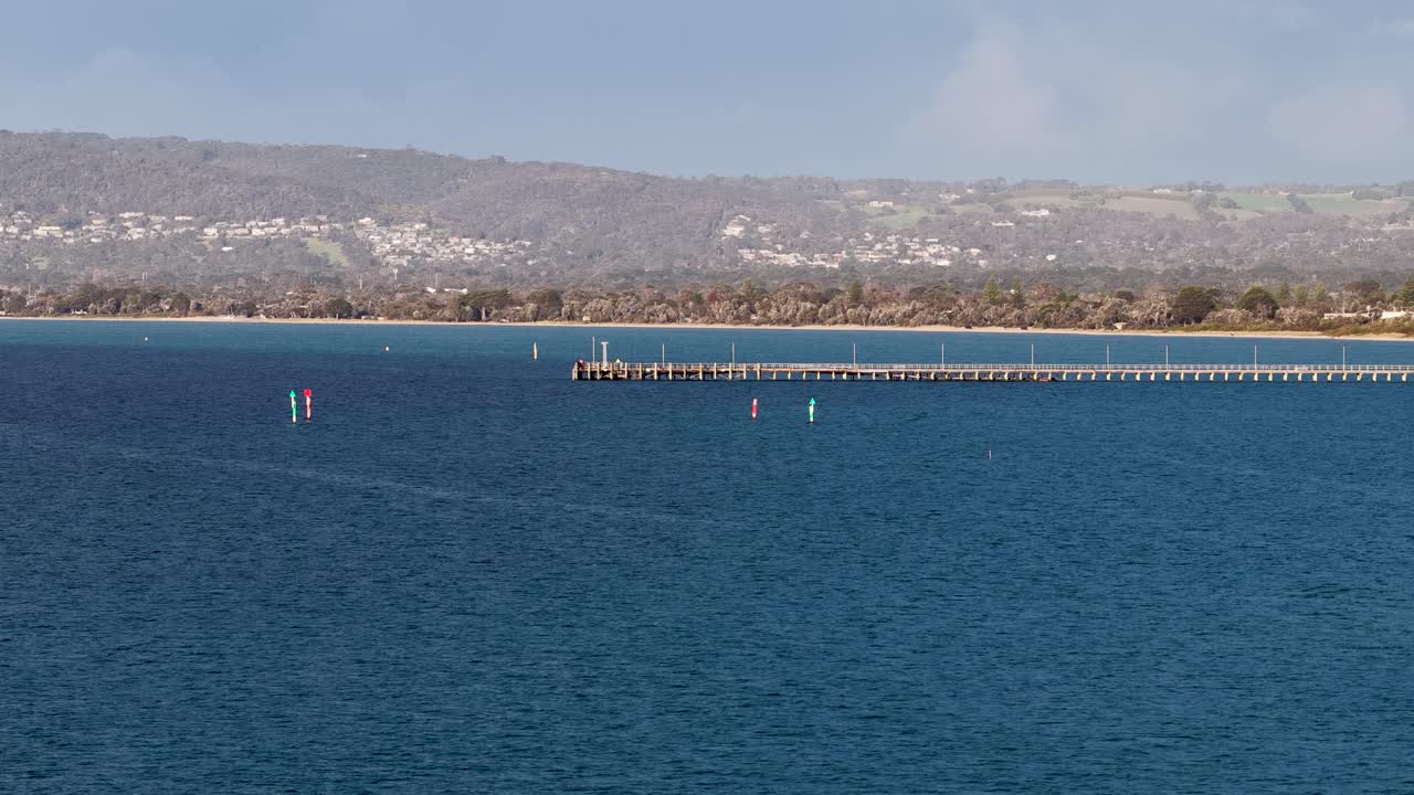 Wide shot of wooden pier stretching into calm bay, bright daylight, static camera, distant hills