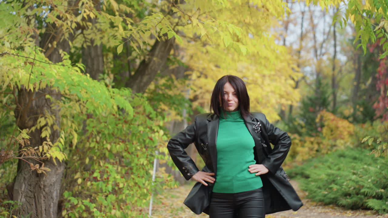 Donna con giacca di pelle nella foresta autunnale, maglione verde e stivali su un sentiero ricoperto di foglie, che cammina e poi si volta prima di guardare la fotocamera, mani sui fianchi in posa rock grintosa, sfondo di fogliame dorato