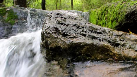 wide angle close up view of a large wet rock with a waterfall behind it to the left and another fully moss covered rock on the right