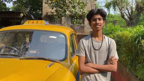 Un-Joven-Taxista-Indio-Con-Una-Camiseta-Gris-Parado-Al-Lado-De-La-Carretera-Al-Lado-Del-Taxi-Y-Sonriendo