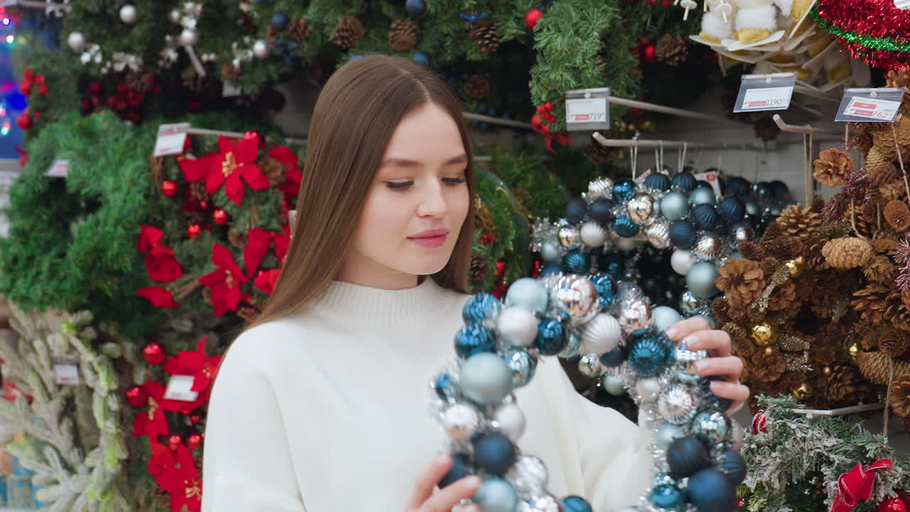 Young woman in white sweater picks up a beautiful Christmas decor wreath and playfully places it on her head while shopping in a festive holiday store surrounded by colorful ornaments