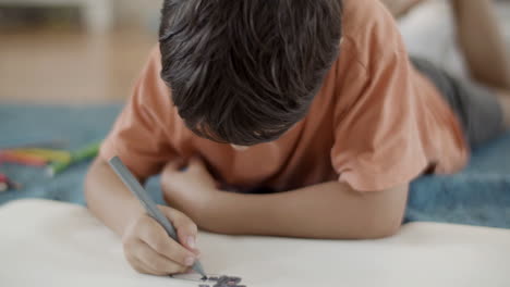 focused little boy lying on floor at home and painting picture