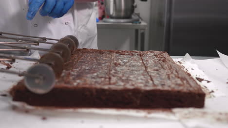 un brownie de chocolate grande es puntuado para cortarlo en múltiples rebanadas usando una rueda de corte de concertina por una chef blanca con guantes azules