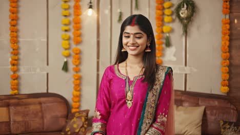 portrait of beautiful young hindu woman wearing traditional pink and green sari and gold jewelry, posing in a decorated home interior with orange flower garlands and sofa