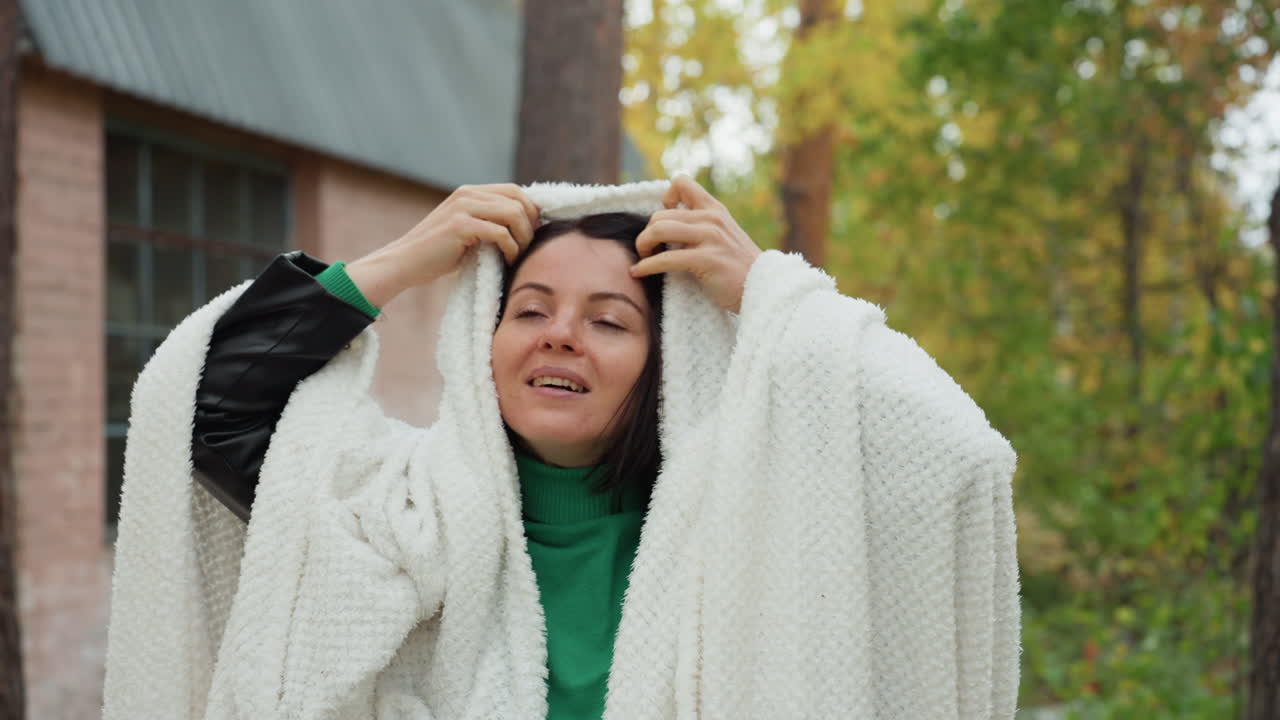 Donna avvolta in un grosso plaid in un cottage. Scena autunnale all'aperto presso un cottage rustico con catasta di legna e porta scolorita, donna con maglione verde che si avvolge uno scaldacollo di lana attorno alle spalle e sistema il cappuccio