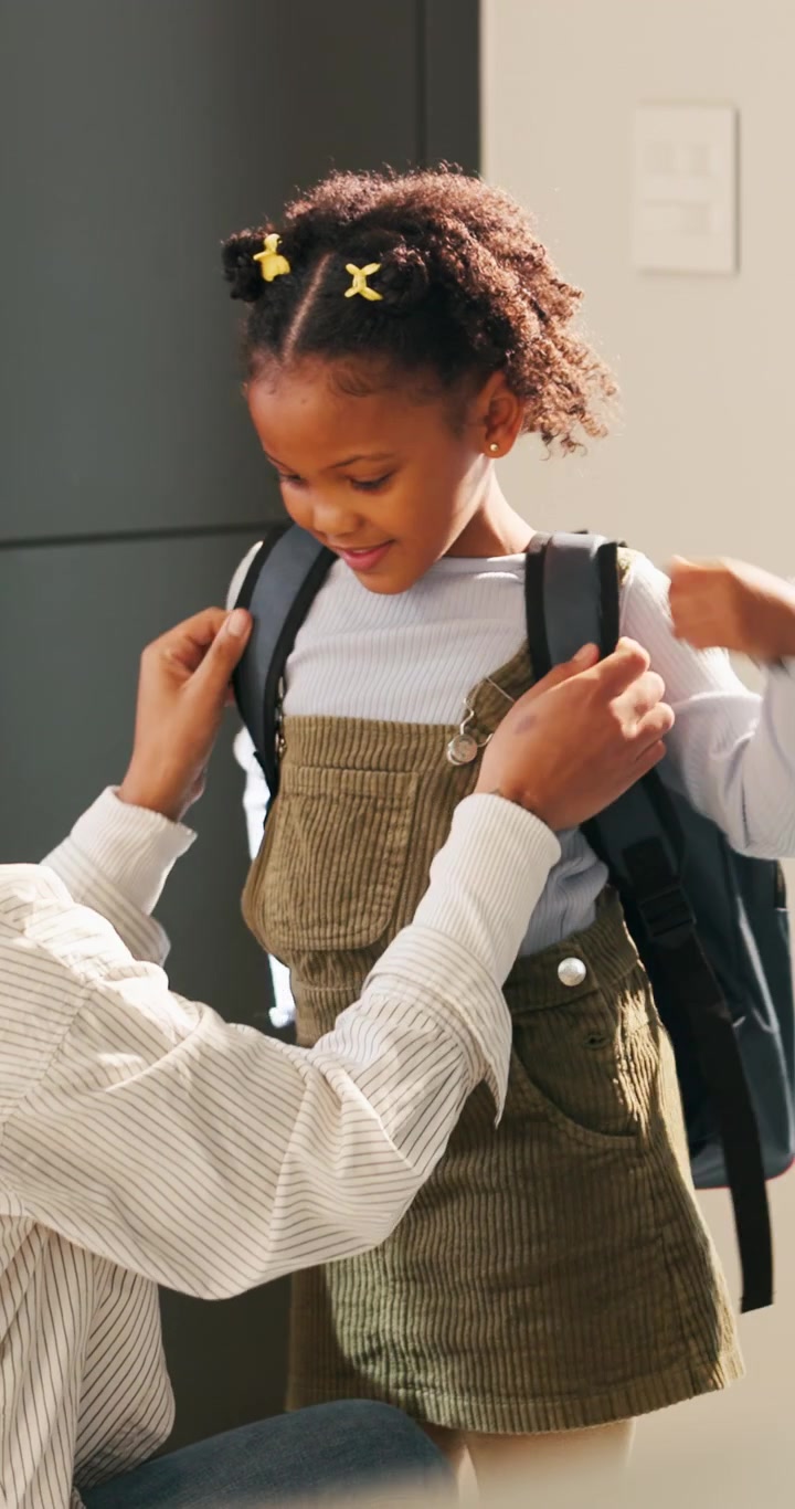 Mother Helping Her Young Daughter Put On A Backpack For School