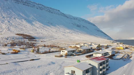 aerial follow back over small snowy winter village at bottom of snow covered mountain hill in westfjords in iceland