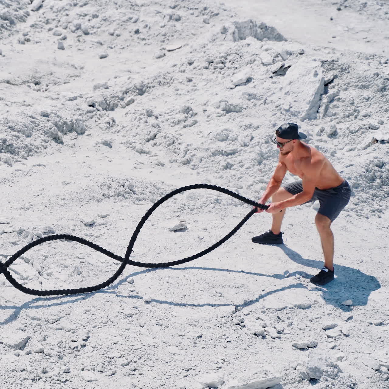 Athletic male performs battle ropes exercise on the white rocky background. Sportsman in shorts doing waves with cable during an outdoor fitness workout.