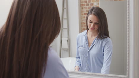 woman wearing pyjamas at home in modern bathroom brushing teeth reflected in mirror