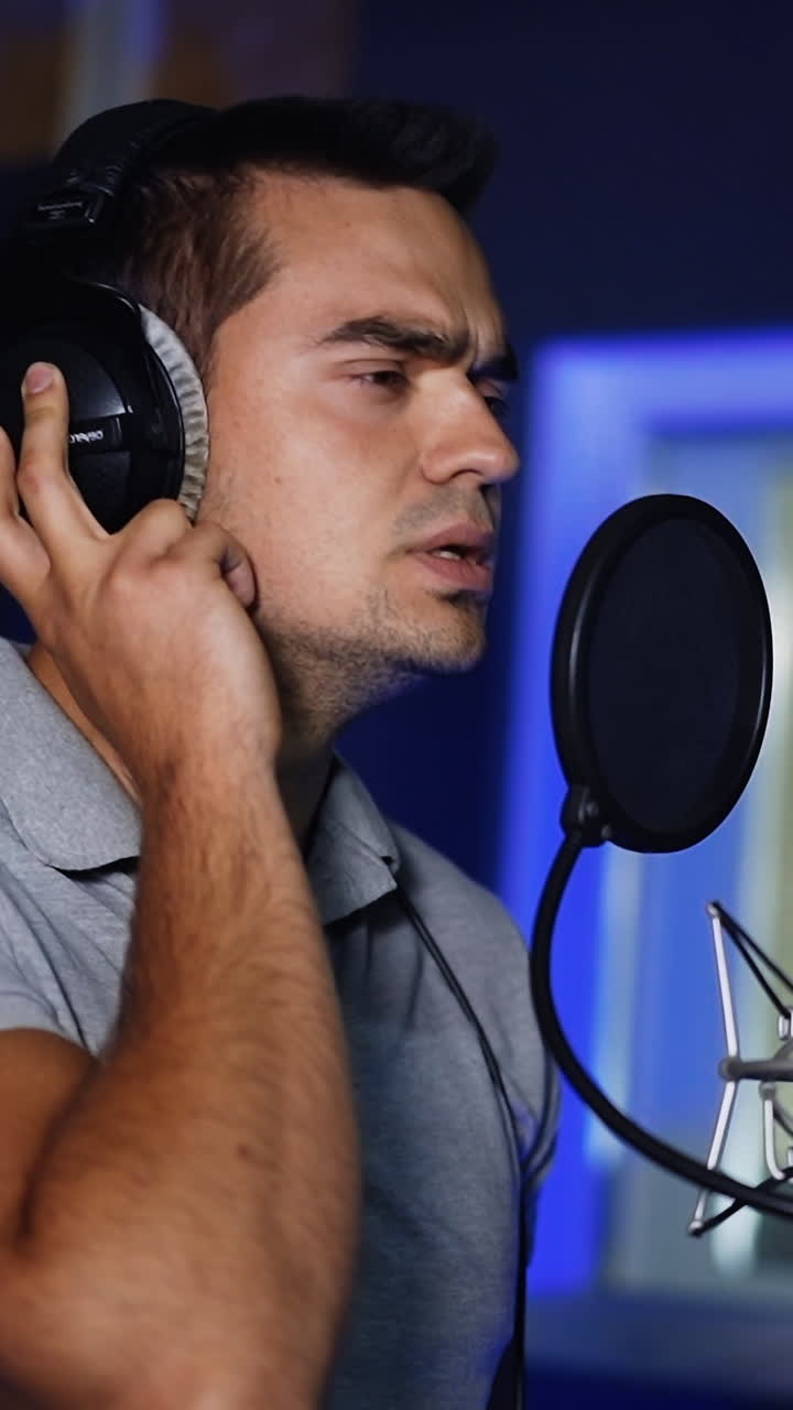 Handsome male wearing headphones singing in studio. Professional singer performing a song, recording music in modern company. Vertical video