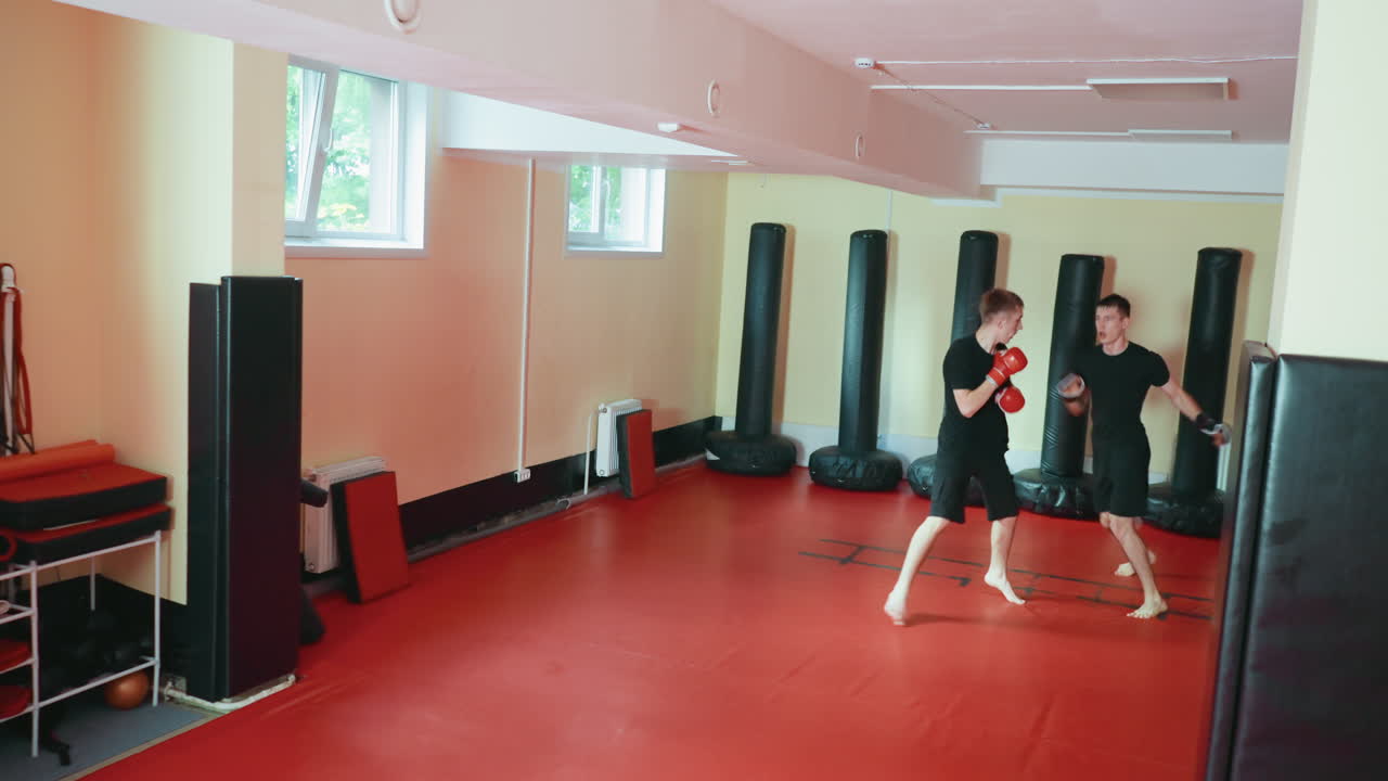 자연광이 드는 martial arts gym에서 한 파이터가 강력한 펀치를 날리며 앞으로 돌진하고, 상대는 빨간색 매트 바닥에서 펀칭백으로 둘러싸인 채 반격하는 가라테 수련생들의 격렬한 스파링