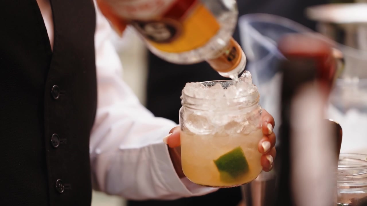 Un cameriere con camicia bianca e gilet scuro porge un bicchiere pieno di ghiaccio tritato, un cocktail chiaro e uno spicchio di lime fresco al bancone bar