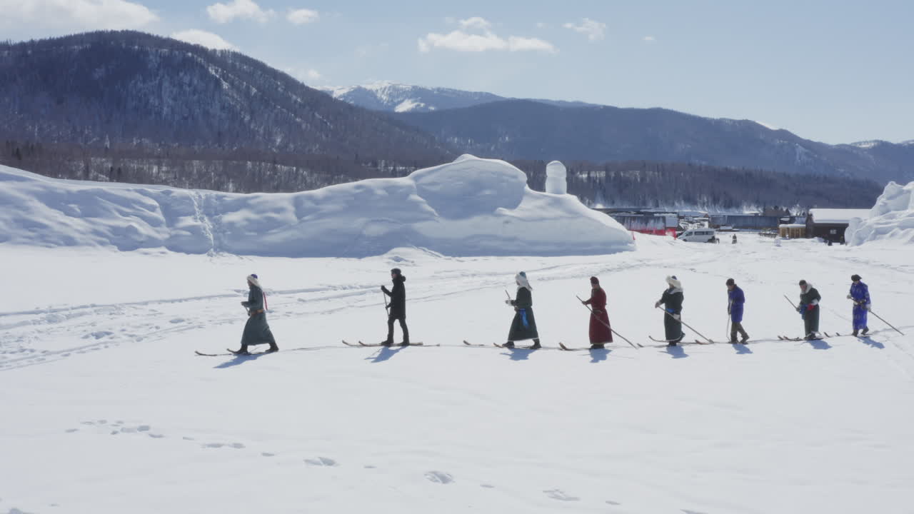 Tuva men dressed in traditional robes walking in snow with fur skis on winter day, aerial epic