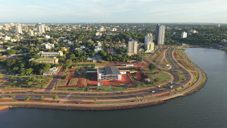 modernist la usina stands near the bay in posadas, argentina, along costanera avenue, surrounded by parks, palm trees, traffic lanes and tall buildings, with river edge in foreground, drone orbiting