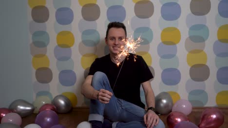 Young-Man-Sitting-with-a-Sparkler-Against-Colorful-Background,-Surrounded-by-Balloons-on-the-Ground