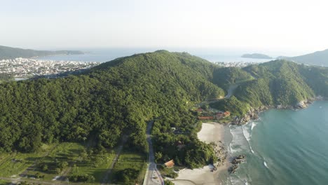 cinematic aerial drone video of jungle mountains with a road revealing a city behind it, bombinhas, santa catarina, brazil