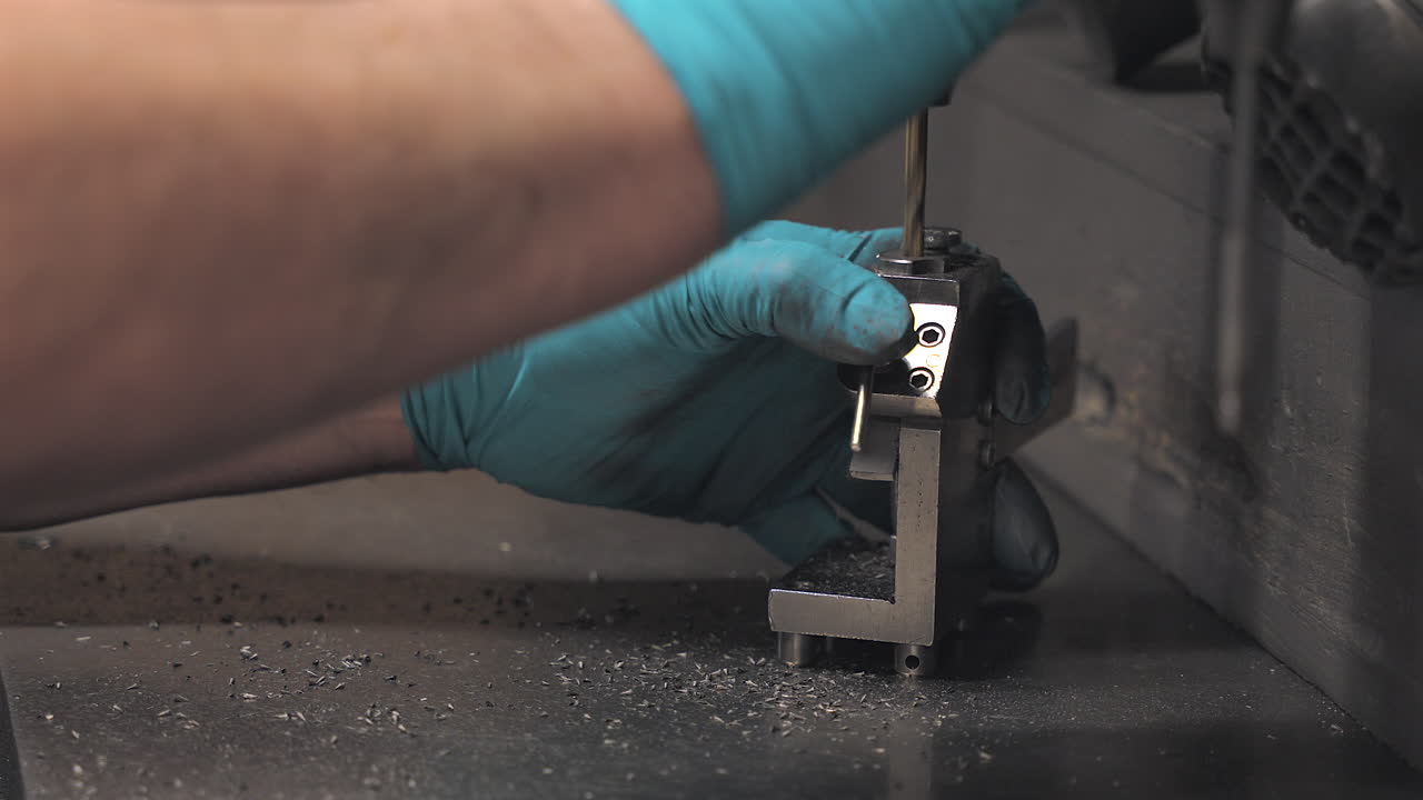 Close-up of a technician using a drill press and fixture to machine a small metal part, showcasing precision craftsmanship in a manufacturing setting.