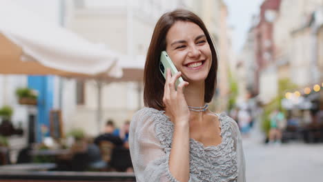 Happy-young-woman-having-remote-conversation-talking-on-smartphone,-good-news-gossip-in-city-street