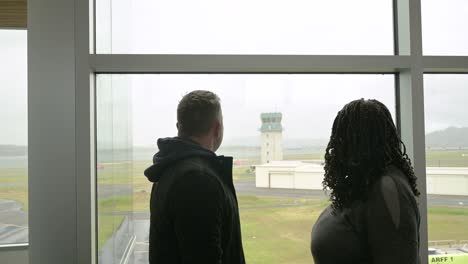 hombre caucásico y mujer negra miran a la torre del aeropuerto por la ventana, luego se besan de despedida.