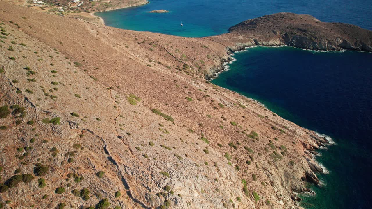 Aerial bird's eye view tilt up along dry coastline of syros greece, reveals sailboat and peninsula