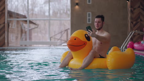 funny man is sitting on inflatable duck in swimming pool and filming himself by smartphone