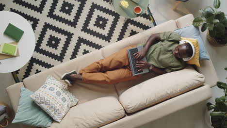 black man in headphones resting on couch and using laptop