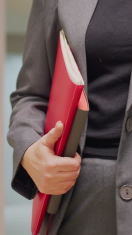 close-up of businesswoman holding red folder in office corridor, dressed in gray suit, confident professional in modern work environment, career-focused, stylish business attire