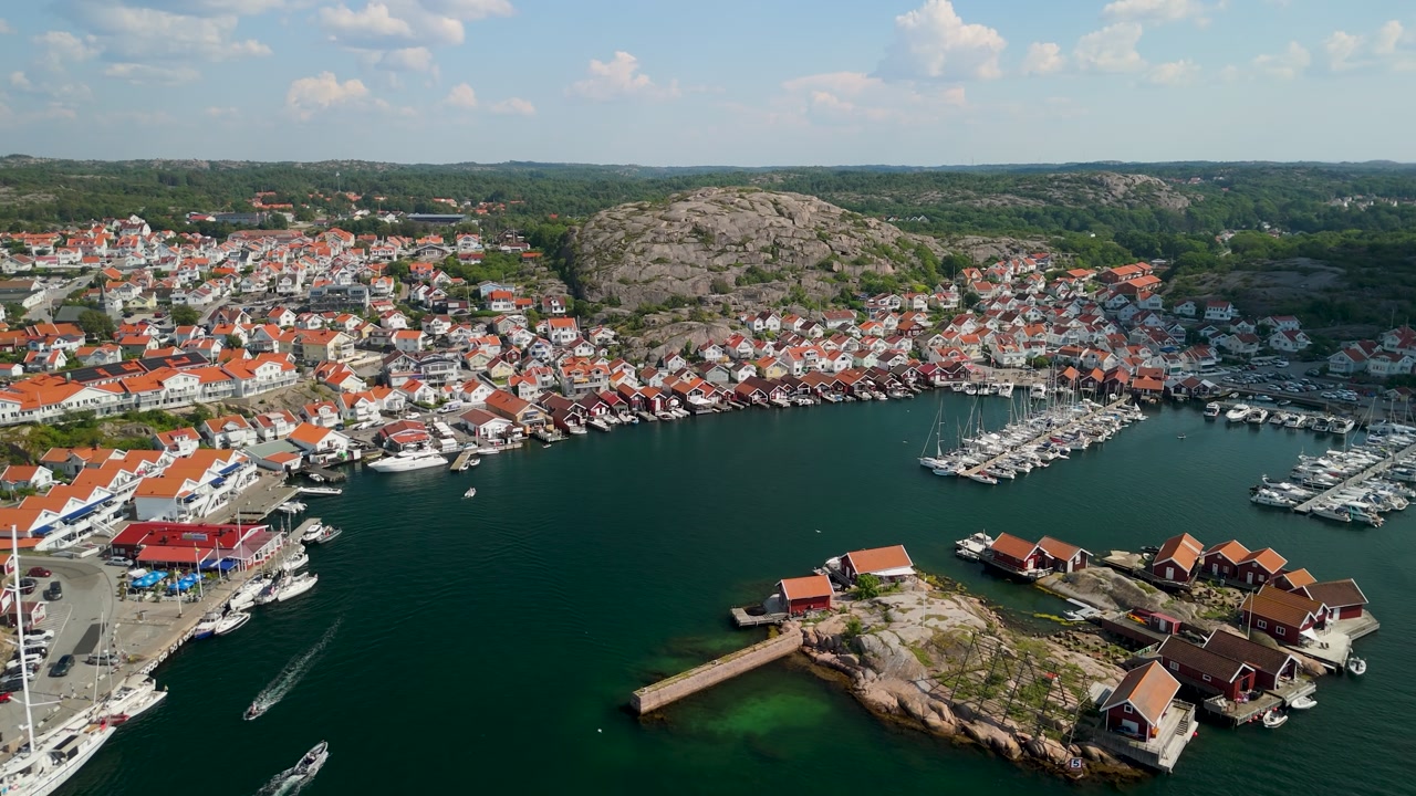 Aerial descent above Hunnebostrand harbor in Bohuslän Sweden revealing dense coastal town wrapping around calm bay with marina sailboats and granite hill