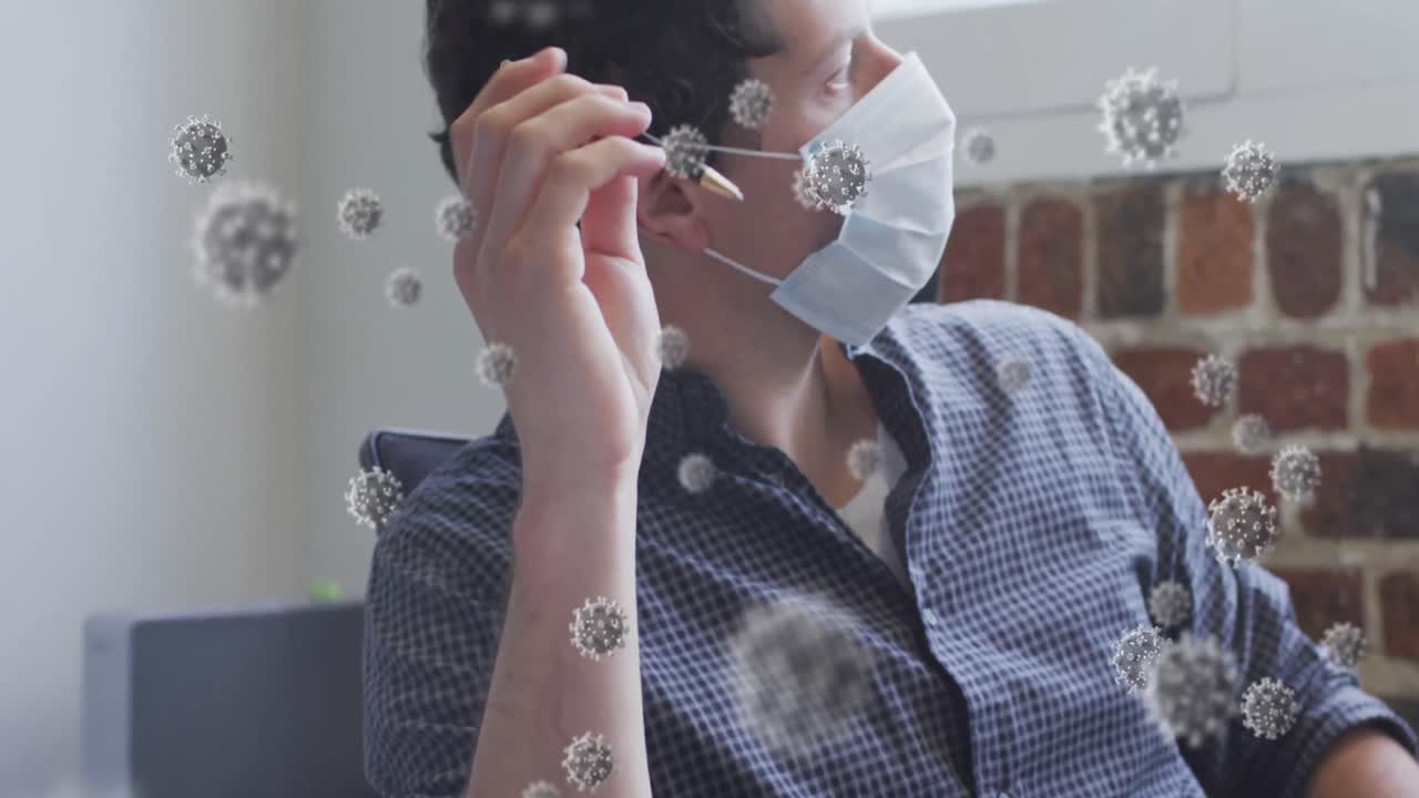 Animation of covid 19 cells floating over man wearing face mask, sitting by desk in office
