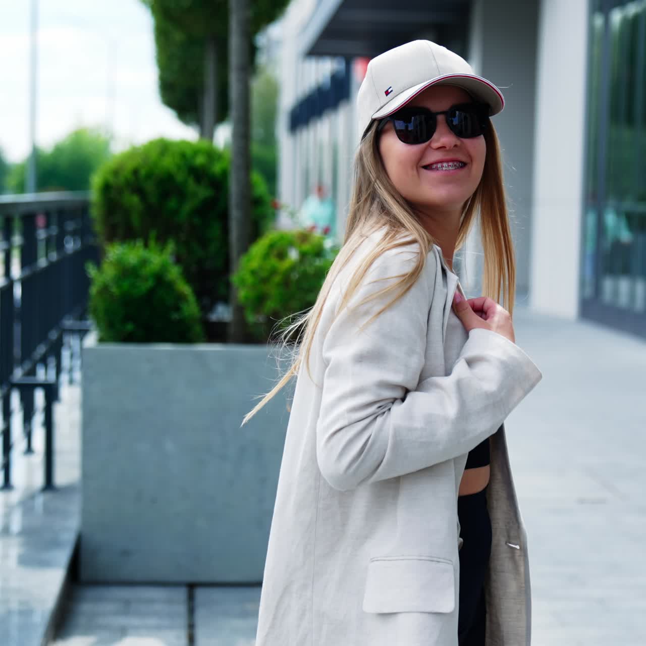 Blonde long-haired model turning in front of camera. Lady in jacket, cap and sunglasses demonstrating clothes in the street. Blurred backdrop