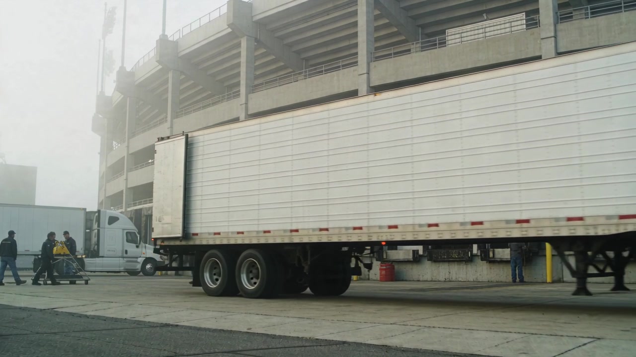 Moving crew pushing cart toward stadium gate after white semi parking, wearing hivis vest for setup