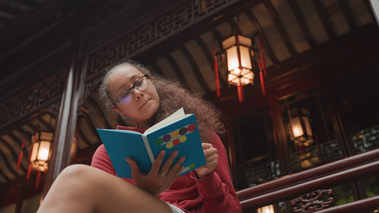 Studente nero che legge un libro in un padiglione ornato, calda luce di lanterna, capelli ricci, occhiali, maglione rosso, balcone di legno, atmosfera serale, espressione concentrata, ritratto cinematografico dal basso