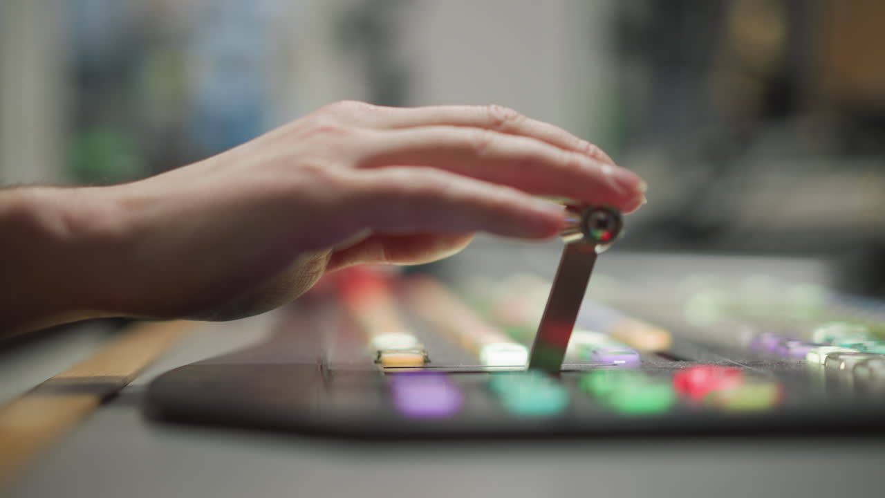 Close-up of hand pressing colorful buttons on broadcast control panel with blurred background, focusing on television production equipment and media control