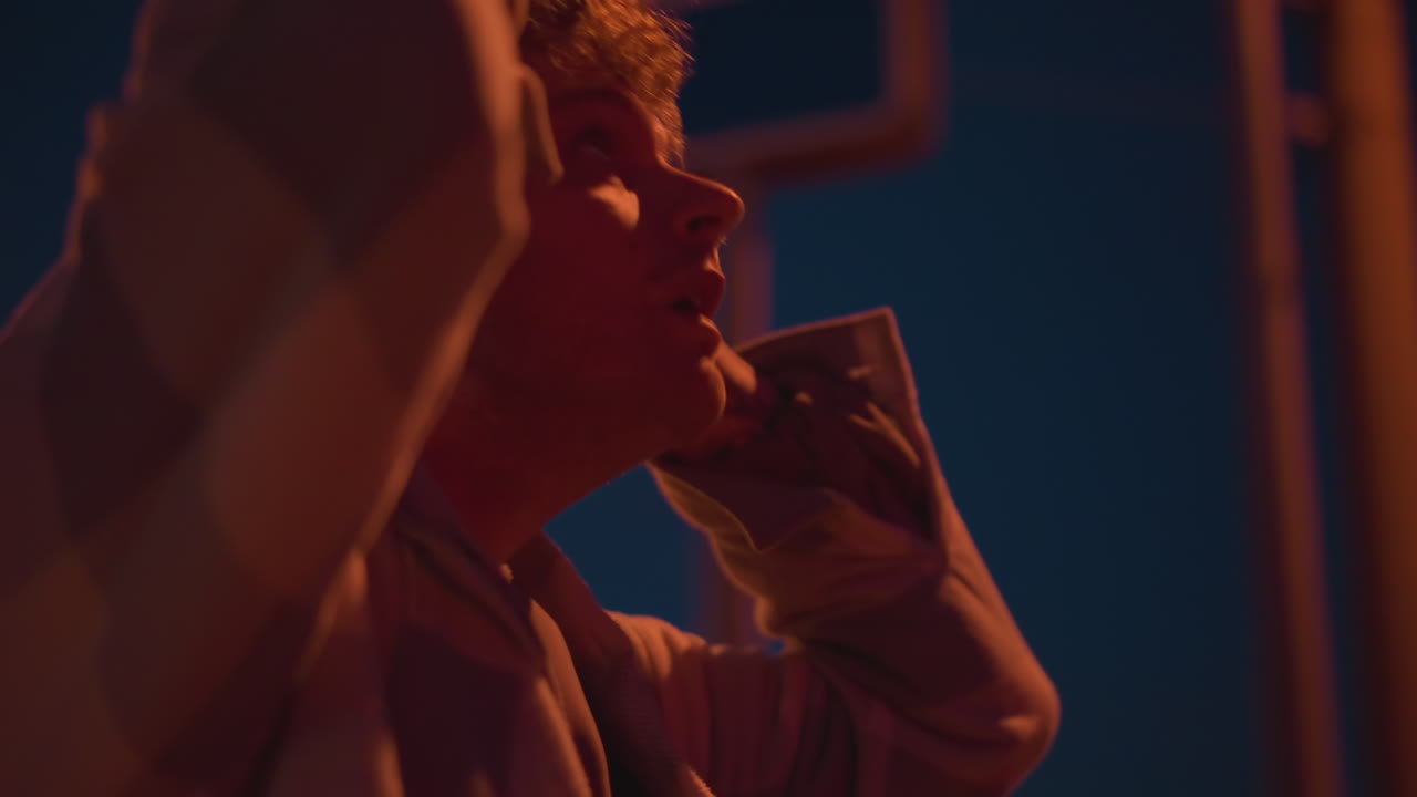 Giovane con felpa con cappuccio notturna che sistema il berretto sotto la soffusa luce di un lampione, primo piano di profilo e movimento delle mani alla testa, espressione introspettiva con calda luce arancione, texture cinematografica suggestiva e viaggio intimo