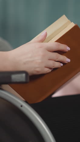 hands of woman in wheelchair flipping pages of book with hard brown cover. female person with spinal cord injury enjoys reading at home close view