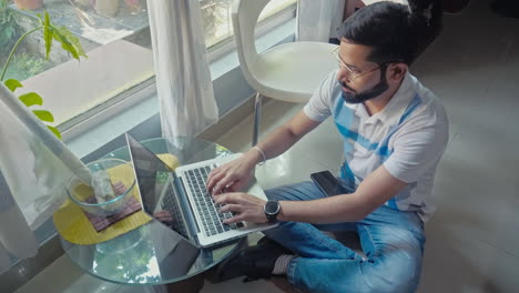 top view of an indian man sitting on the floor, holding mobile phone and using laptop computer near window side.
