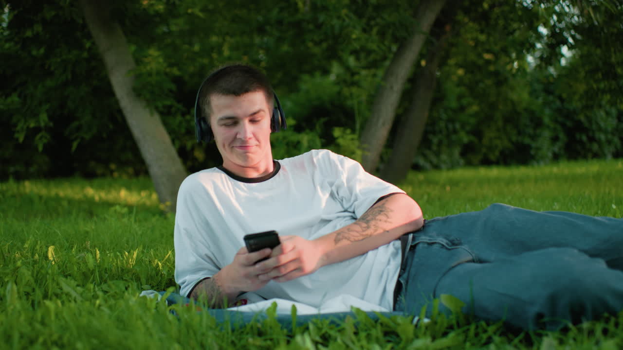 헤드폰을 착용한 원격 노동자가 손에 드폰을 들고 잔디에 누워서 음악에 고개를 숙이고 미묘한 미소로 나무와 활기찬 초록색으로 둘러싸여 부드러운 빛 아래 있습니다.