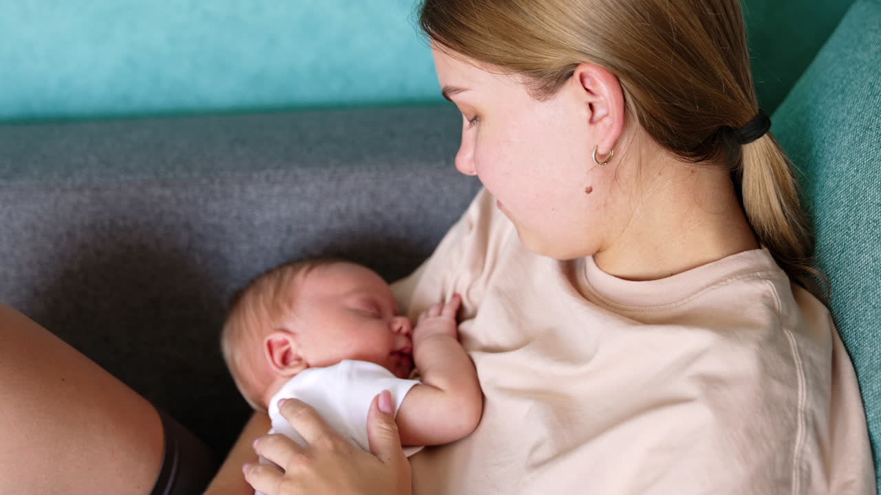 Blond Caucasian woman holding her sleeping newborn. Mom touches her child's ear tenderly. Close up.