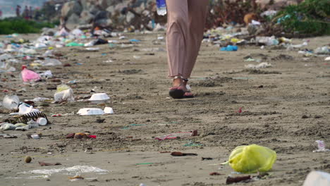 una mujer caminando sobre la arena rodeada de basura mientras sostiene una botella de agua, tiro estático