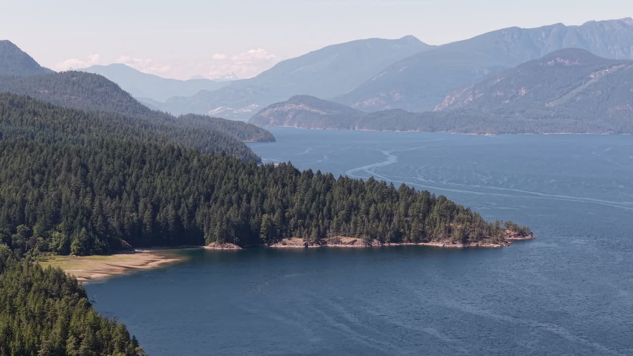 A 70mm lens drone video of a tree covered rocky point that meets the ocean. Beyond the point are layers of west coast mountains!