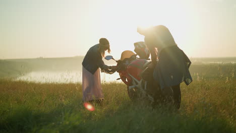 two women are cleaning a red motorcycle in a grassy field with the sun reflecting on the other lady, one woman is bent over the bike, and the other is carefully wiping the front