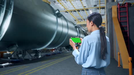 vista trasera de una mujer de negocios asiática usando un teléfono móvil con una pantalla verde simulada en una fábrica de fabricación de tubos