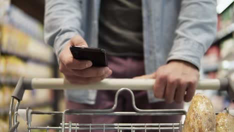 Cropped-close-up-footage-of-a-young-man-pushing-trolley-cart-against-blurred-background-and-shopping-in-grocery-supermarket.-Concentrated-male-using-modern-smartphone-and-choosing-fresh-food