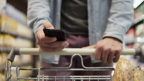 cropped close up footage of a young man pushing trolley cart against blurred background and shopping in grocery supermarket. concentrated male using modern smartphone and choosing fresh food