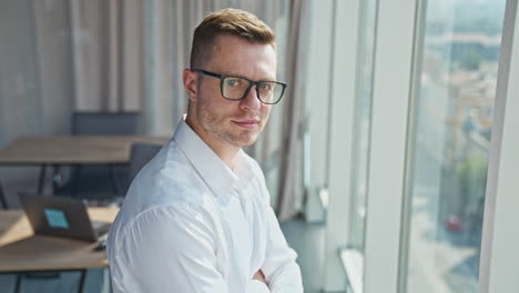 confident handsome mid-aged man wearing glasses looks into camera. businessman standing in the office near a big panoramic window in the rays of sun.