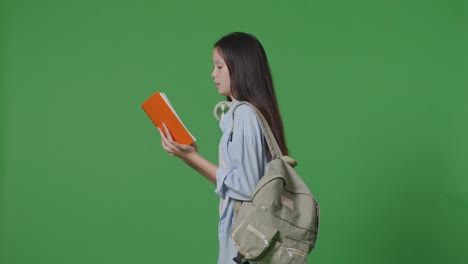 side view of asian teen girl student with a backpack holding and reading a book while walking in the green screen background studio