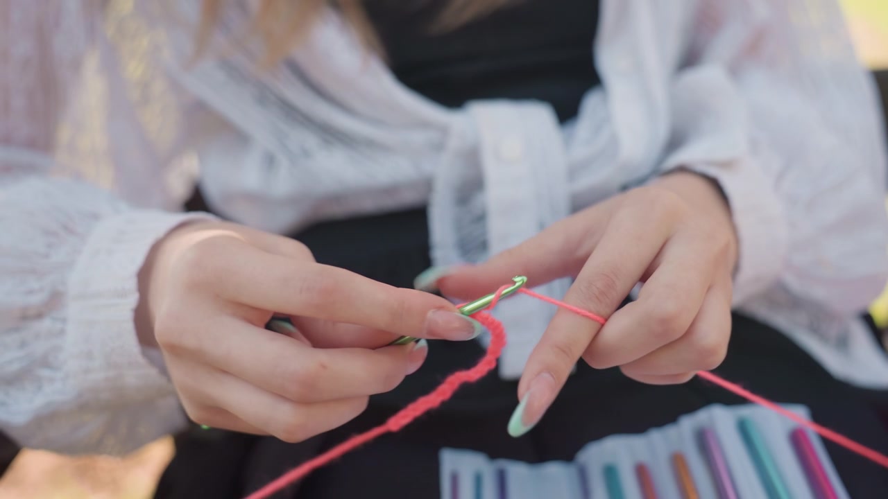 Primo piano di una giovane donna che lavora all'uncinetto con filato rosa e un uncinetto verde, con le mani a fuoco. La donna indossa una blusa di pizzo bianco sopra un top nero e ha diversi uncinetti colorati visibili in grembo. Ambiente esterno con luce soffusa.