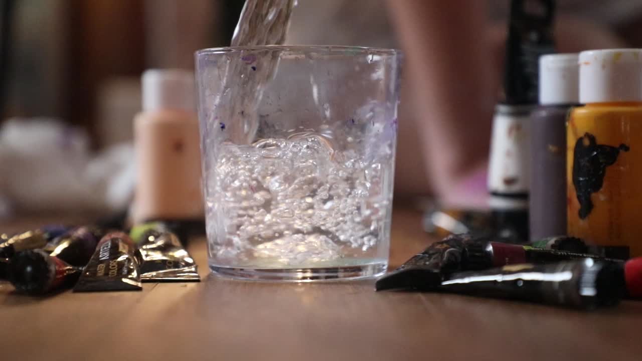 Slow-motion of water poured in glass for painting purposes. Camera moves away from the glass as the water is being poured, painting tubes and instruments.